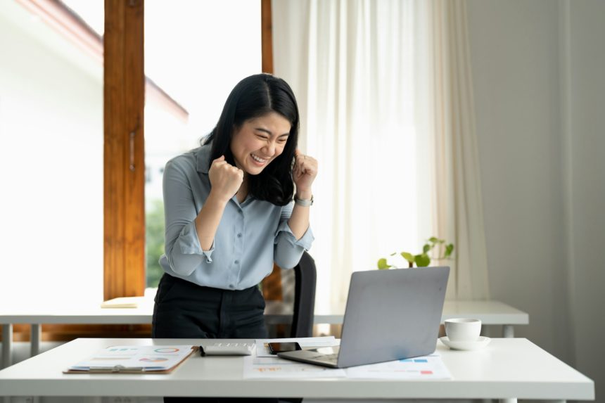 Excited young woman standing at table with laptop and celebrating success. achieving goals excited