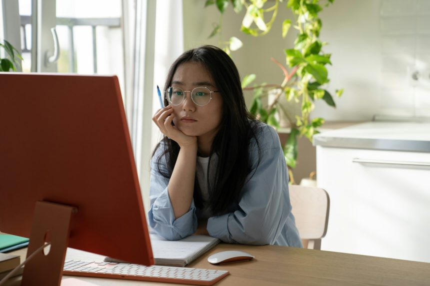 Young focused Asian girl student watching webinar video course on computer during online learning